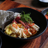 Noodle dish with vegetables and red chili peppers in a black bowl on a wooden surface