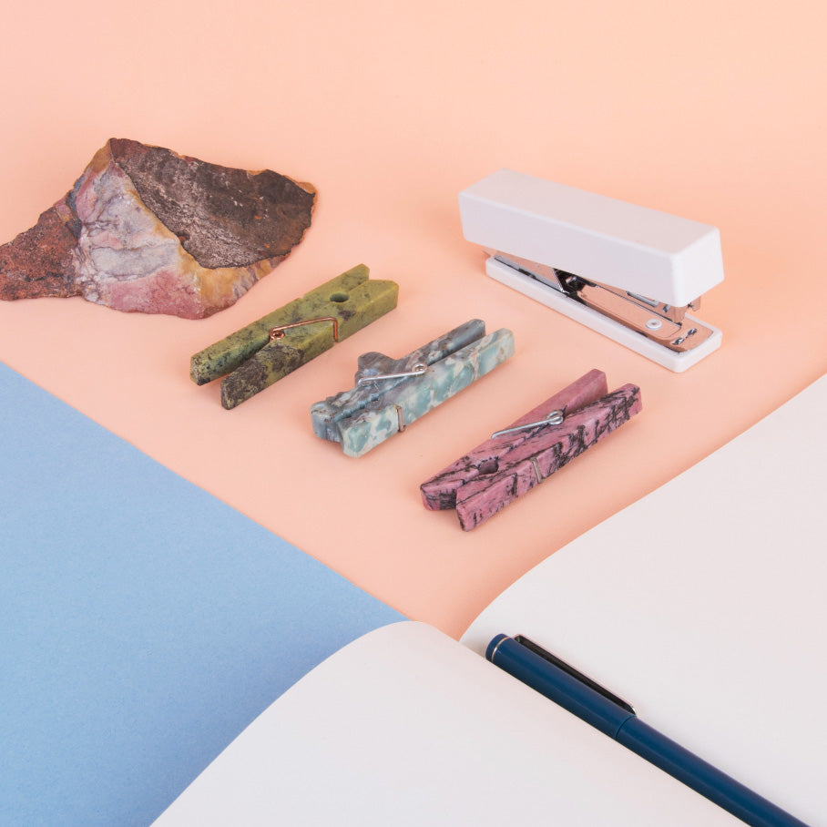 Decorative stone clothespins next to a notebook and stapler on a pink and blue background