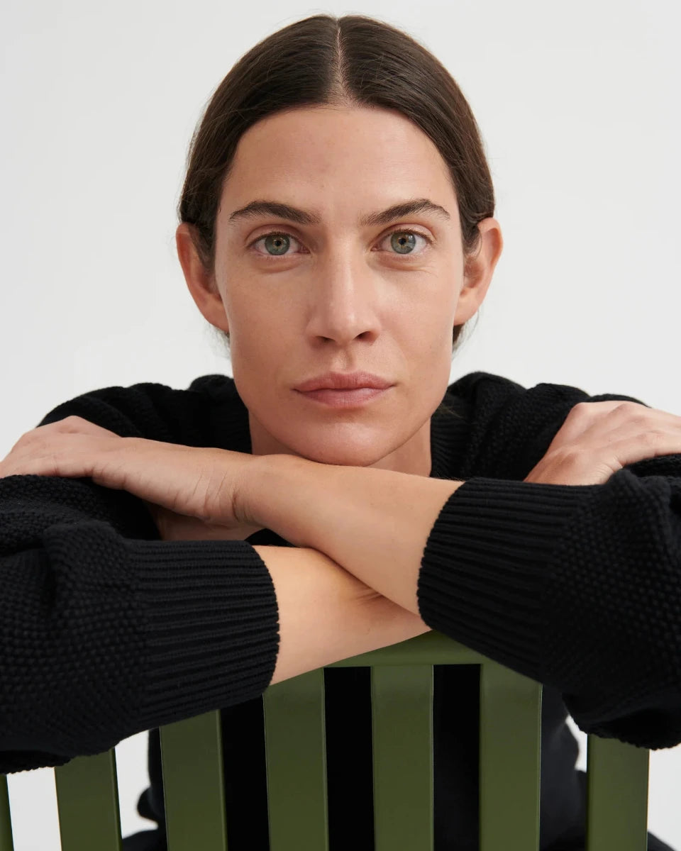 A close-up portrait of a woman with dark hair and green eyes, wearing a black ribbed knit sweater and resting her arms on her hands, leaning over a green slatted chair against a light gray background.