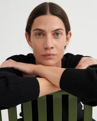 A close-up portrait of a woman with dark hair and green eyes, wearing a black ribbed knit sweater and resting her arms on her hands, leaning over a green slatted chair against a light gray background.