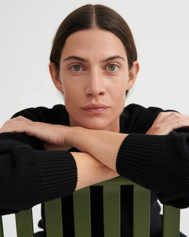 A close-up portrait of a woman with dark hair and green eyes, wearing a black ribbed knit sweater and resting her arms on her hands, leaning over a green slatted chair against a light gray background.