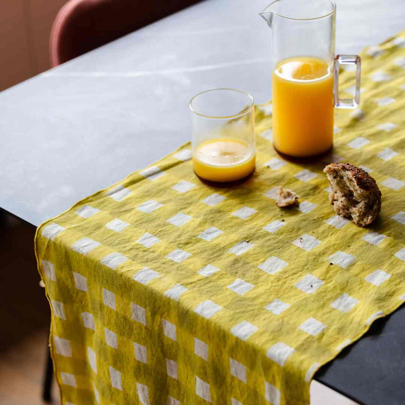Glass of orange juice and pitcher on a yellow checkered table runner with a piece of bread.
