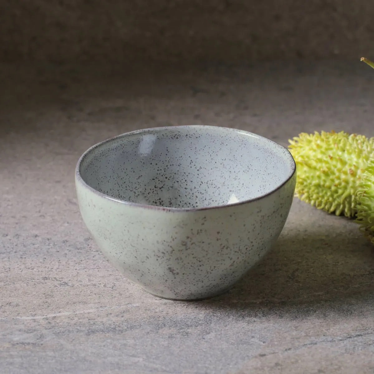 Speckled gray ceramic bowl on a textured surface with a green fruit in the background