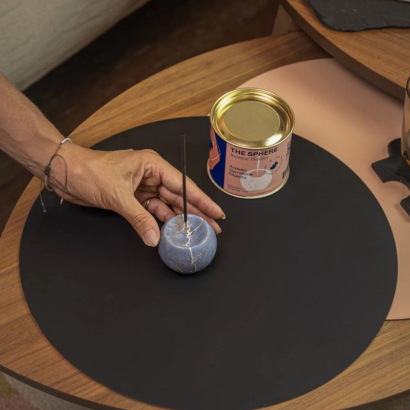 Hand interacting with a small blue sphere incense holder on a black mat next to a can labeled 'The Sphere'.