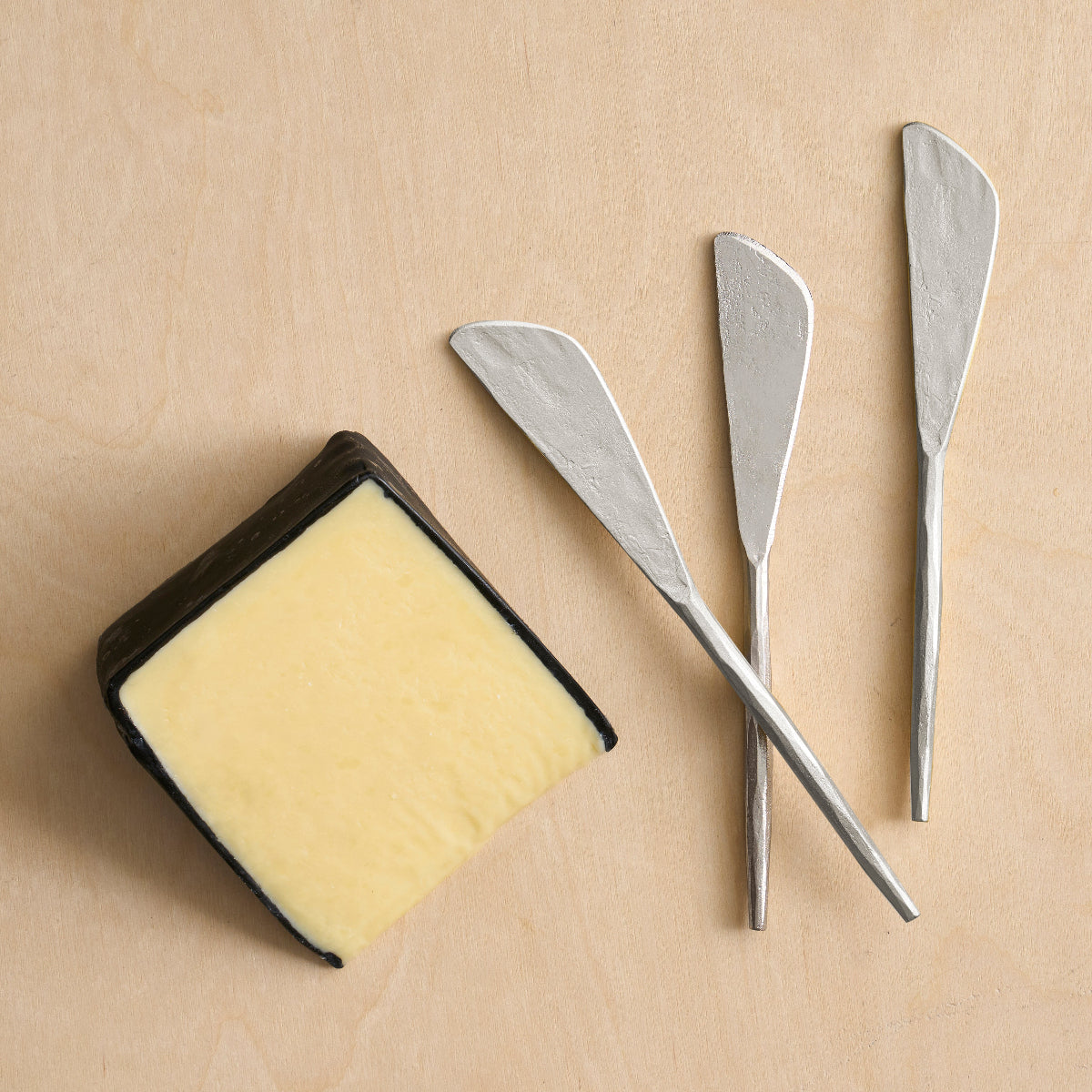 Three pewter spreader knives and a slice of cheese on a wooden surface