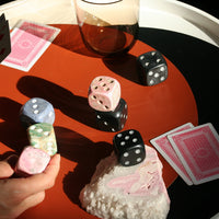 an assortment of black and colorful stone dice on a surface with cards, a glass, and a crystal object