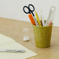 Green cup holding stationery items on a wooden surface with an hourglass in the background
