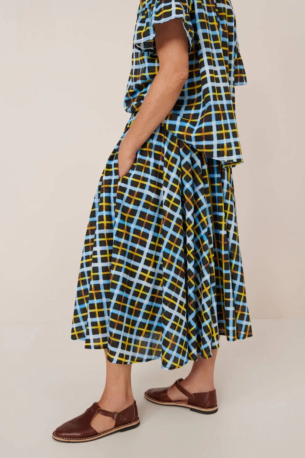 Person wearing a blue and yellow plaid shirt and skirt with brown sandals on a beige background