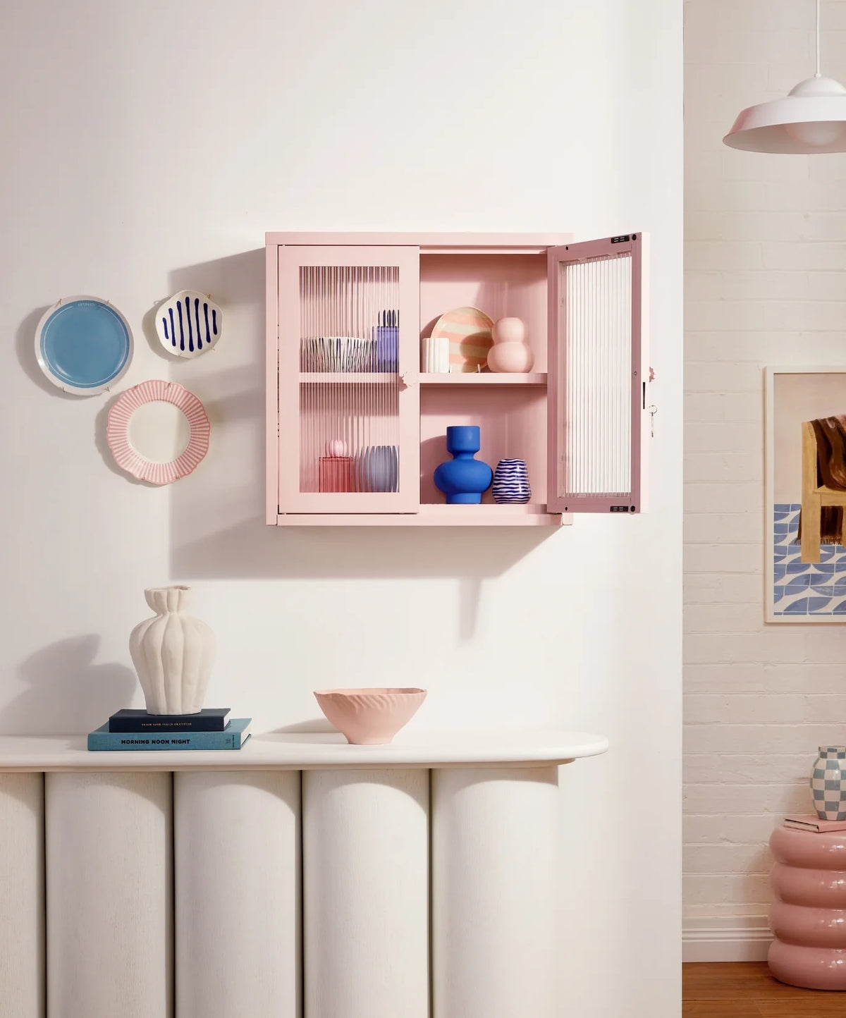 Pink cabinet with decorative items on a white wall with a small table and vases.