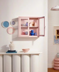 Pink cabinet with decorative items on a white wall with a small table and vases.