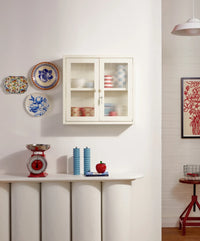 White cabinet with glass doors, decorative plates, and a red scale on a white wall.