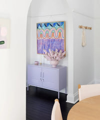 Light purple sideboard with a vase of pink flowers and abstract painting on a white wall.