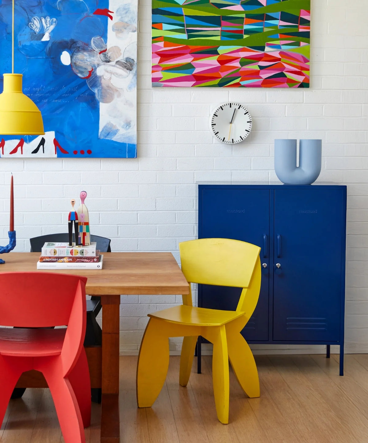 Colorful dining area with red and yellow chairs, a blue cabinet, and abstract art on the wall.