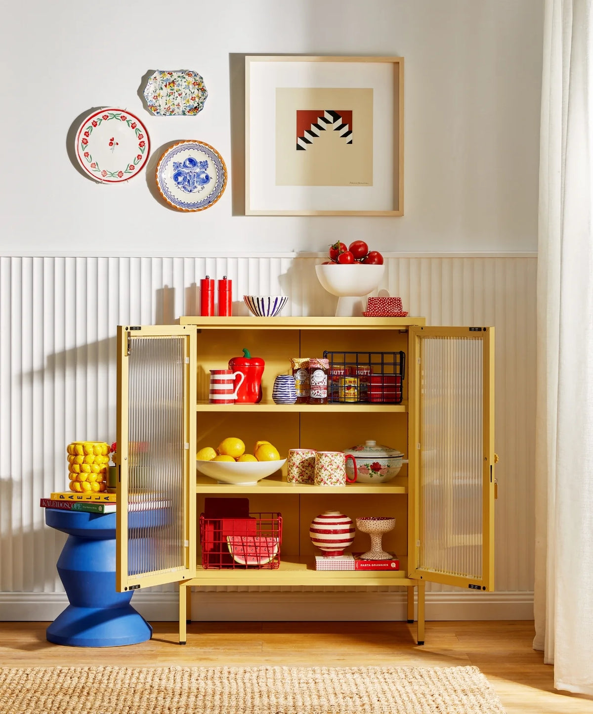 Yellow cabinet with open doors displaying various items in a room with decorative plates and a painting on the wall.