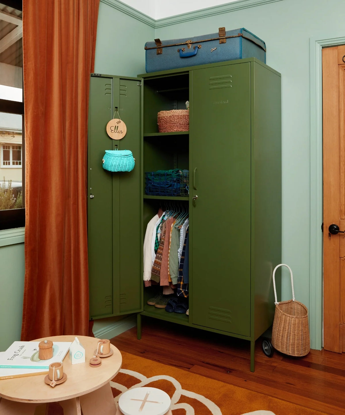 Green metal wardrobe with clothes and items in a room with wooden floor and light blue walls.