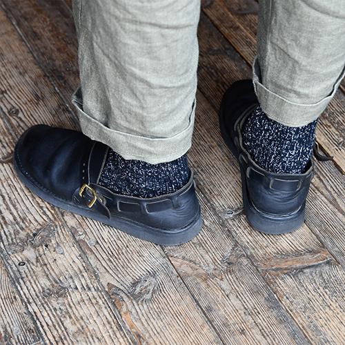 back of person's ankles wearing beige linen pants, speckled navy socks, and dark brown leather clogs on a wooden surface