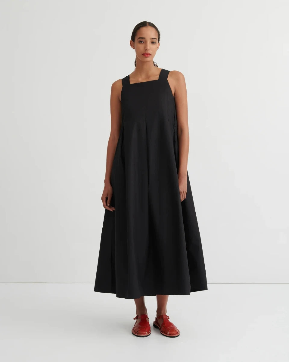 A woman stands against a plain light gray studio background wearing a sleeveless black midi dress with a square neckline and a flared skirt, paired with red shoes.