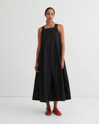 A woman stands against a plain light gray studio background wearing a sleeveless black midi dress with a square neckline and a flared skirt, paired with red shoes.