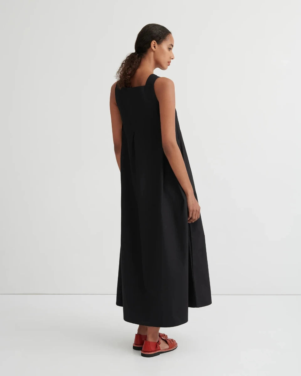 A woman wearing a sleeveless black midi dress, shown from a rear three-quarter view against a plain light gray studio background.