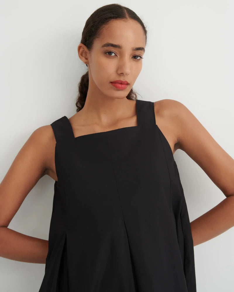 A woman wearing a black sleeveless top with a square neckline and wide shoulder straps, posed against a plain light gray background.