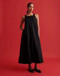 A woman modeling a sleeveless black midi dress with a square neckline and side pockets, standing against a solid red studio background.
