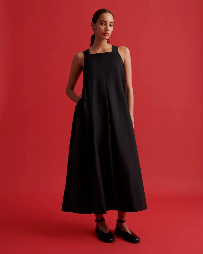A woman modeling a sleeveless black midi dress with a square neckline and side pockets, standing against a solid red studio background.