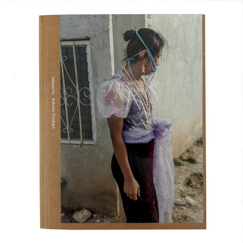 A square book titled TRÁNSITO by Roberto Tondopó against a white background. The cover features a photographic scene of a person standing outdoors beside a weathered wall, wearing a sheer white lace top, dark red skirt, turquoise beaded headpiece, and a light purple wrap. 