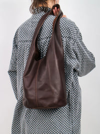 Person with a polka dot shirt wearing a dark brown leather bag over one shoulder against a plain background