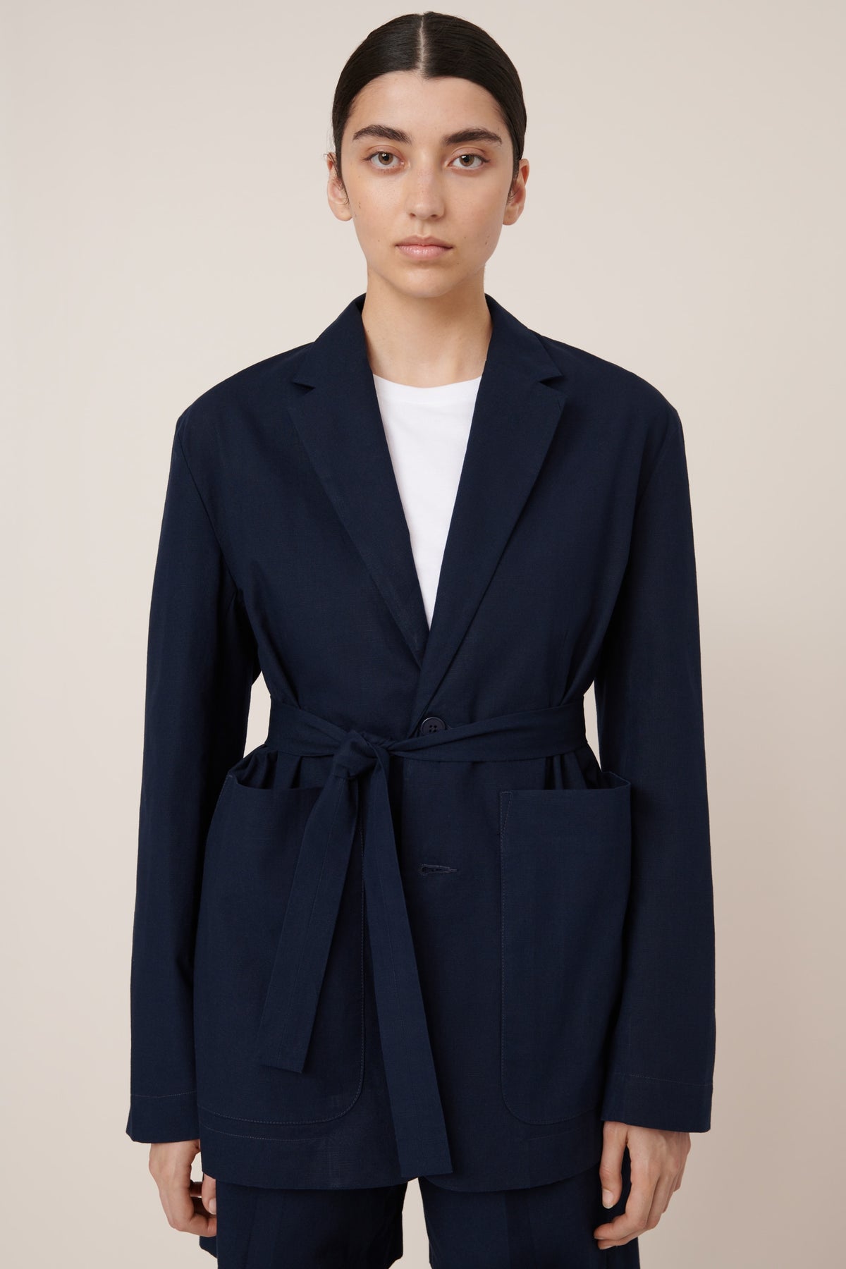 Person wearing a belted navy blue coat with a white shirt underneath on a beige background