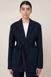 Person wearing a belted navy blue coat with a white shirt underneath on a beige background