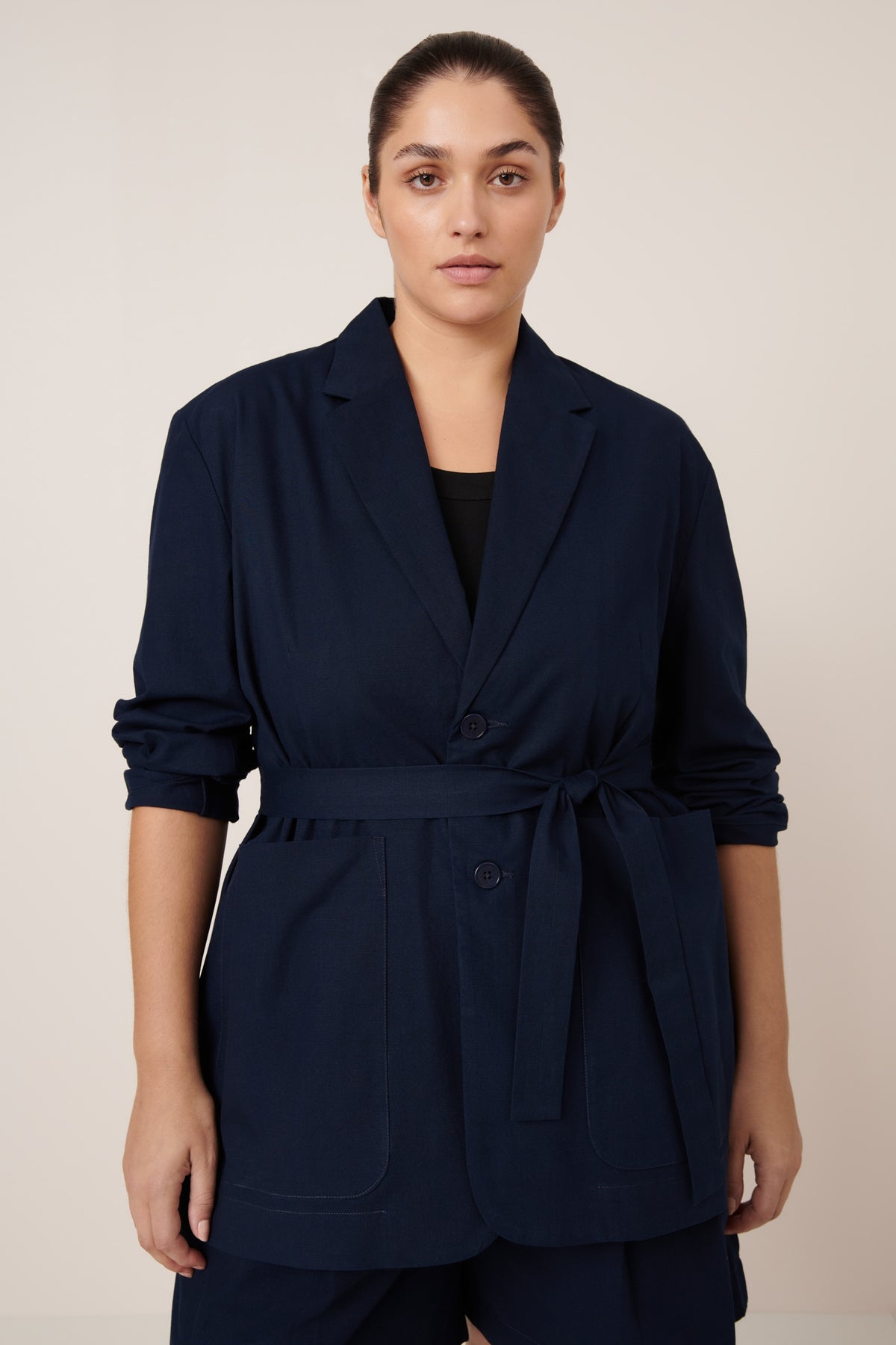 Woman wearing a navy blue belted jacket with rolled up sleeves on a beige background